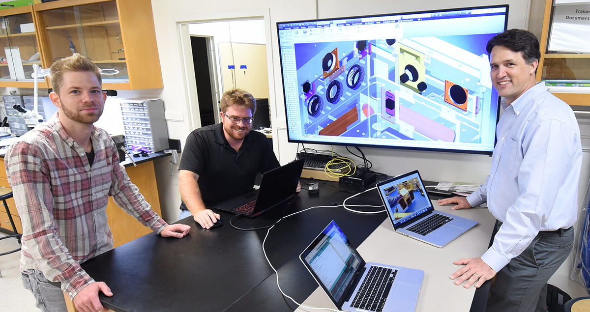 Two unamed male grad students and Dr. Gunter, all standing astride a computer screen displaying their cubesat
