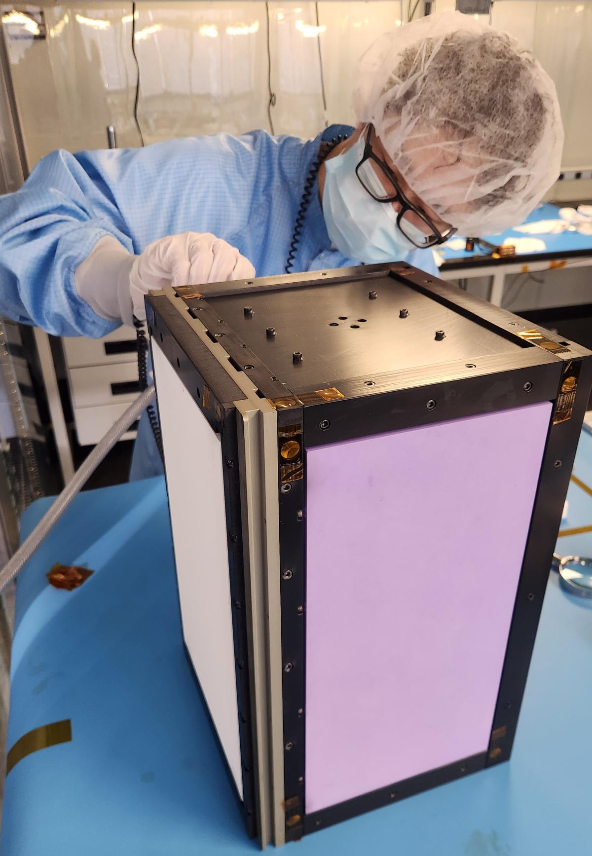 A Georgia Tech student in a cleanroom suit and mask inspects a CubeSat named OrCa2